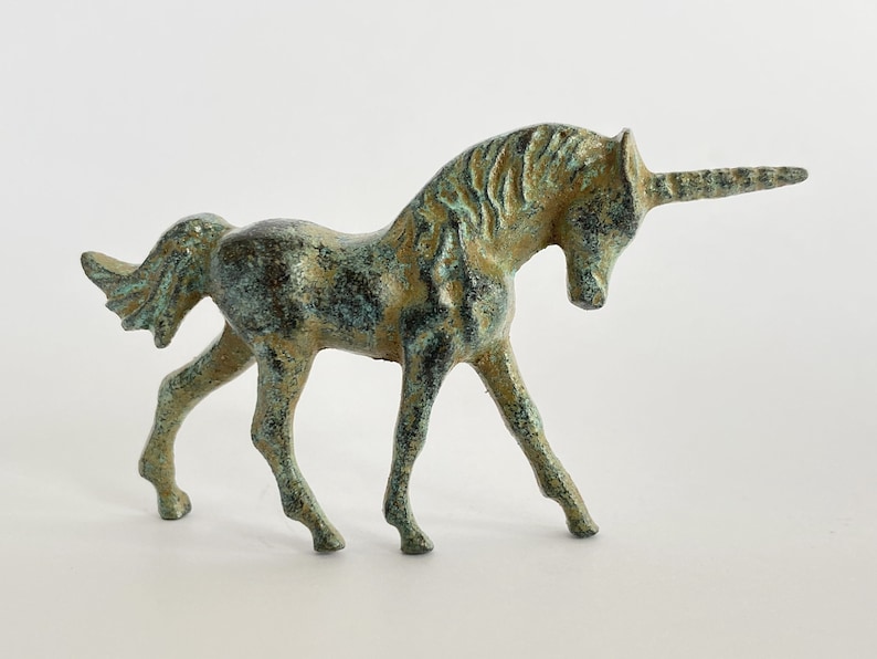 May include: A small, green and gold metal unicorn figurine. The unicorn is standing on all four legs with its head turned to the side. The unicorn has a long, thin horn.