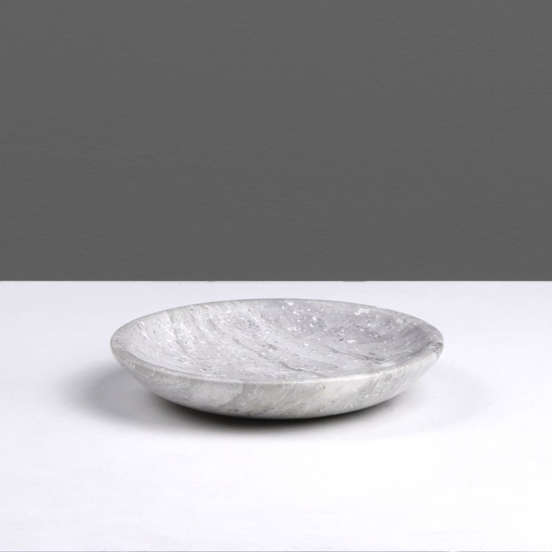 May include: A round, gray marble dish with a slightly rippled surface. The dish is empty and sitting on a white surface.