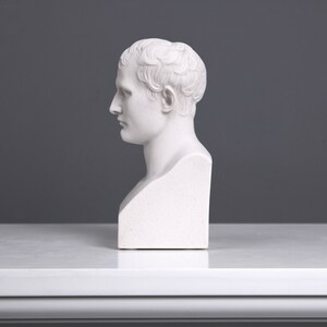 May include: White plaster bust sculpture of a man with short hair. The bust is on a white pedestal.