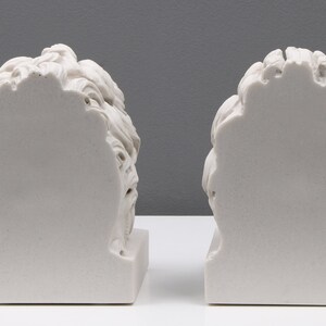 Może przedstawiać: Dwa białe marmurowe popiersia os&oacute;b z plecami do widza. Popiersia stoją na białym tle.