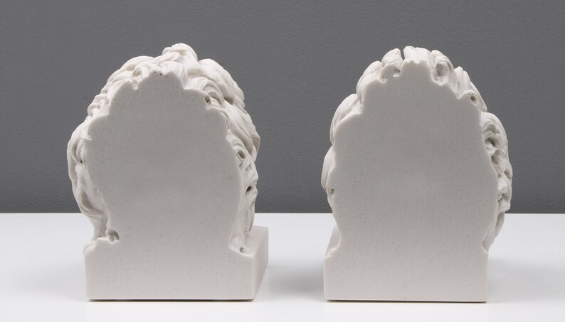 Może przedstawiać: Dwa białe marmurowe popiersia os&oacute;b z plecami do widza. Popiersia stoją na białym tle.