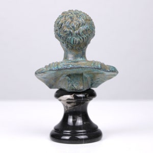 May include: A small, green bronze bust of a person on a black marble base. The bust is of a person with short hair and is facing forward.