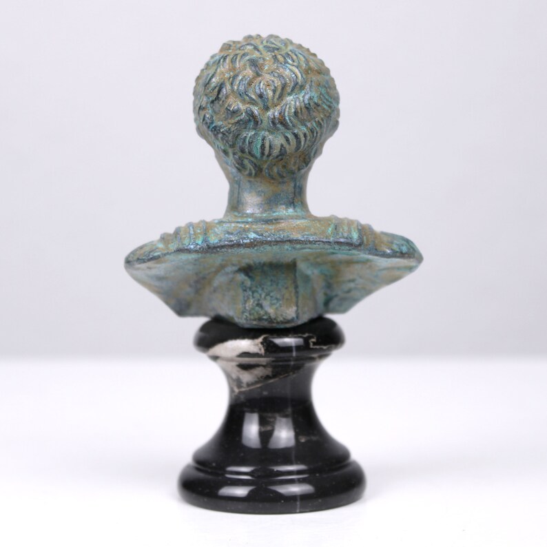 May include: A small, green bronze bust of a person on a black marble base. The bust is of a person with short hair and is facing forward.