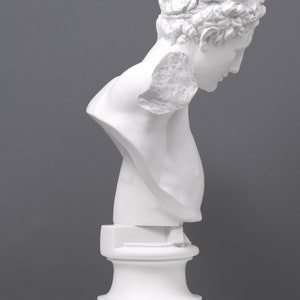 May include: White plaster bust sculpture of a person with their head tilted down. The bust is on a white pedestal.