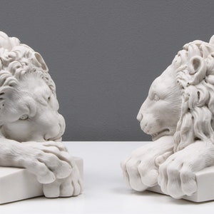 Może przedstawiać: Dwie białe marmurowe rzeźby lw&oacute;w, każda z detaliczną grzywą i spoczywająca na kwadratowej podstawie. Lwy patrzą na siebie, z głowami pochylonymi w d&oacute;ł i łapami spoczywającymi na podstawie.