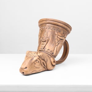 May include: A brown ceramic mug shaped like a ram's head with a decorative design of figures and a torch. The mug has a handle and a spout.