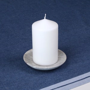 May include: A white pillar candle sits on a gray stone candle holder. The candle and holder are on a blue denim tablecloth.