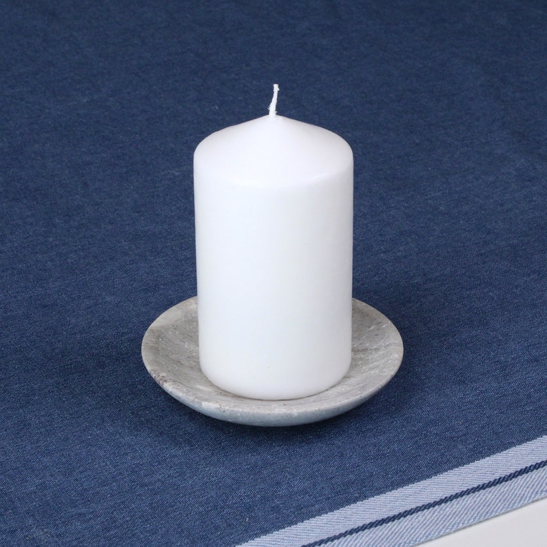 May include: A white pillar candle sits on a gray stone candle holder. The candle and holder are on a blue denim tablecloth.