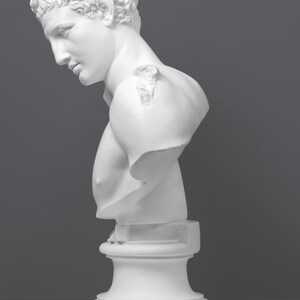 May include: White plaster bust sculpture of a man with short curly hair. The bust is on a white pedestal.