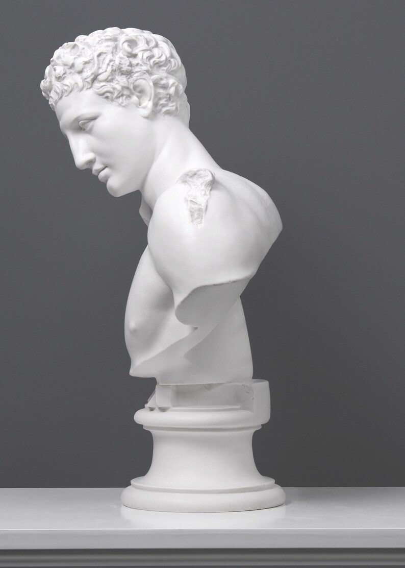 May include: White plaster bust sculpture of a man with short curly hair. The bust is on a white pedestal.