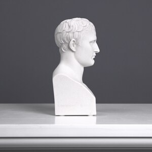 May include: White plaster bust sculpture of a man with short hair. The bust is viewed from the side, showing the man's profile. The bust is on a white surface.