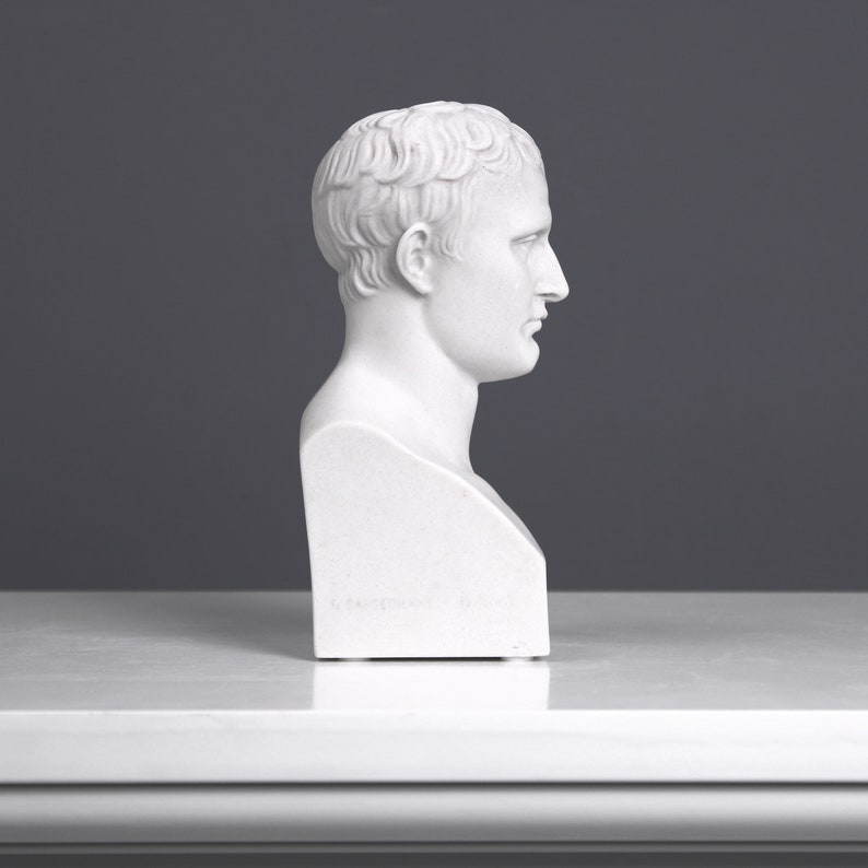 May include: White plaster bust sculpture of a man with short hair. The bust is viewed from the side, showing the man's profile. The bust is on a white surface.