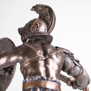 May include: A bronze statue of a gladiator holding a shield and a sword. The gladiator is wearing a helmet with a plume and armor. The statue is detailed and realistic.