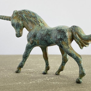 May include: A small, green bronze unicorn figurine with a detailed mane and tail. The unicorn is standing on a light brown surface.