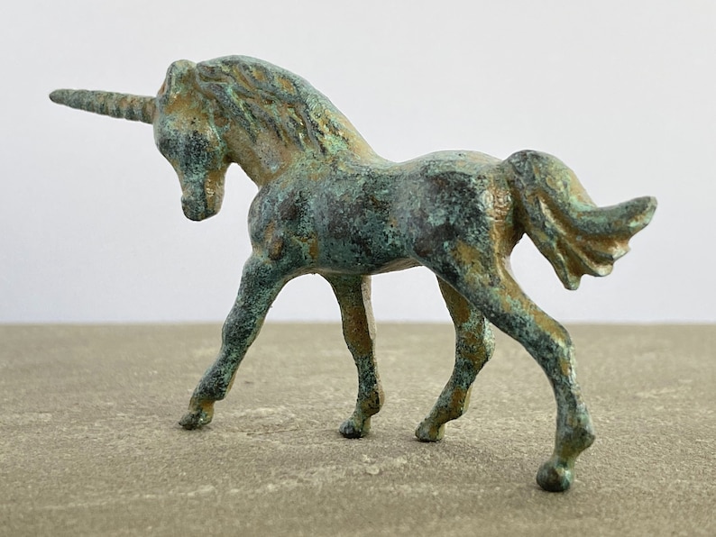 May include: A small, green bronze unicorn figurine with a detailed mane and tail. The unicorn is standing on a light brown surface.