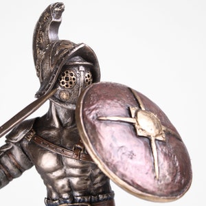 May include: A bronze statue of a gladiator holding a sword and shield. The gladiator is wearing a helmet with a face mask and a leather strap across his chest. The shield is round and has a bronze star in the center.
