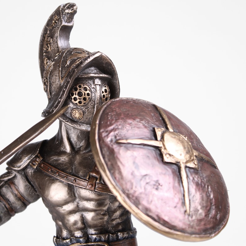 May include: A bronze statue of a gladiator holding a sword and shield. The gladiator is wearing a helmet with a face mask and a leather strap across his chest. The shield is round and has a bronze star in the center.