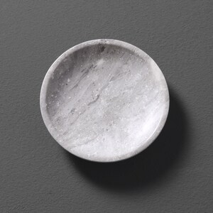 May include: A round, white marble dish with a speckled pattern. The dish is perfect for serving small snacks or holding jewelry.