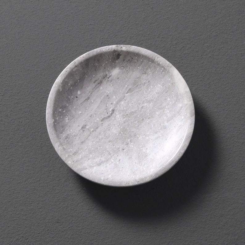 May include: A round, white marble dish with a speckled pattern. The dish is perfect for serving small snacks or holding jewelry.