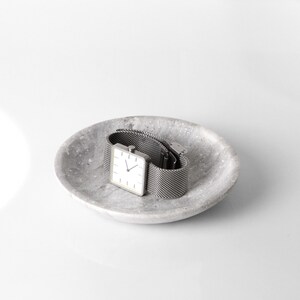 May include: A silver square watch with a white face and a metal mesh band sits on a gray speckled dish.