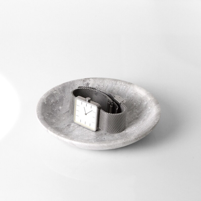 May include: A silver square watch with a white face and a metal mesh band sits on a gray speckled dish.