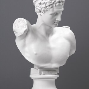 May include: White plaster bust sculpture of a man with short curly hair. The bust is on a white pedestal with the word "Hermes" inscribed on the front.