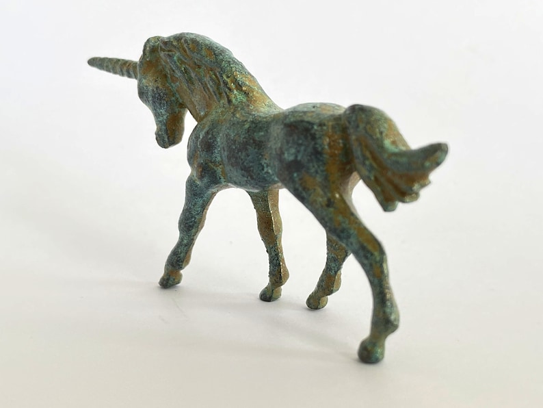 May include: A small, green bronze unicorn figurine with a curled tail and a single horn. The figurine is standing on a white surface.