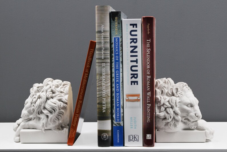 Może przedstawiać: Para białych lwich podp&oacute;r do książek, kt&oacute;re trzymają stos książek. Książki dotyczą sztuki i historii, w tym "Mozaiki świata greckiego i rzymskiego", "Meble" i "Blask rzymskiego malarstwa ściennego".