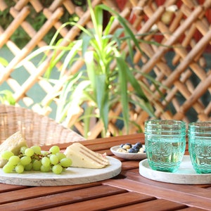 May include: A white marble serving board with a bunch of green grapes, two slices of cheese, and a small bowl of blueberries on a wooden table. Two teal glass cups with decorative designs are on a white marble coaster.