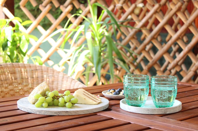 May include: A white marble serving board with a bunch of green grapes, two slices of cheese, and a small bowl of blueberries on a wooden table. Two teal glass cups with decorative designs are on a white marble coaster.