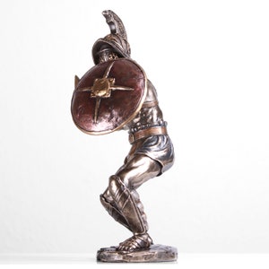 May include: Bronze statue of a gladiator in a fighting pose, holding a shield and sword. The gladiator is wearing armor and a helmet. The statue is on a base.