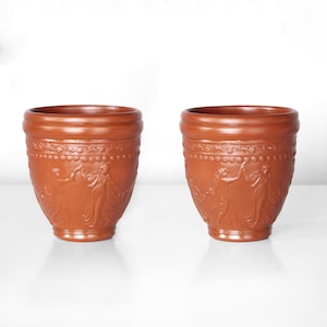 Peut inclure: Deux tasses en terre cuite avec un motif en relief de personnages dans une scène. Les tasses sont brunes et ont une surface lisse et légèrement texturée.