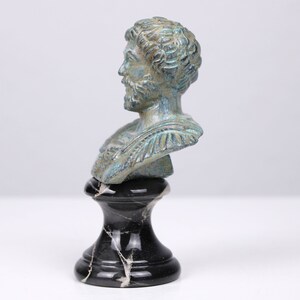 May include: A bronze bust of a man with a beard, on a black marble base. The bust is green and blue, with a patina finish.