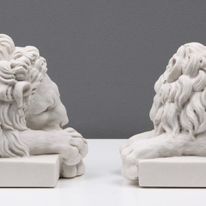 Może przedstawiać: Para białych marmurowych podp&oacute;r na książki w kształcie lwa. Każdy lew leży z głową opartą o łapy. Podpory na książki są szczeg&oacute;łowo wykonane z realistycznym futrem i rysami twarzy.