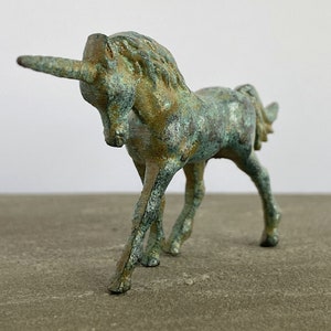 May include: A small, green and gold metal unicorn figurine. The unicorn is standing on all four legs with its head turned to the side. The unicorn has a long, curved horn.