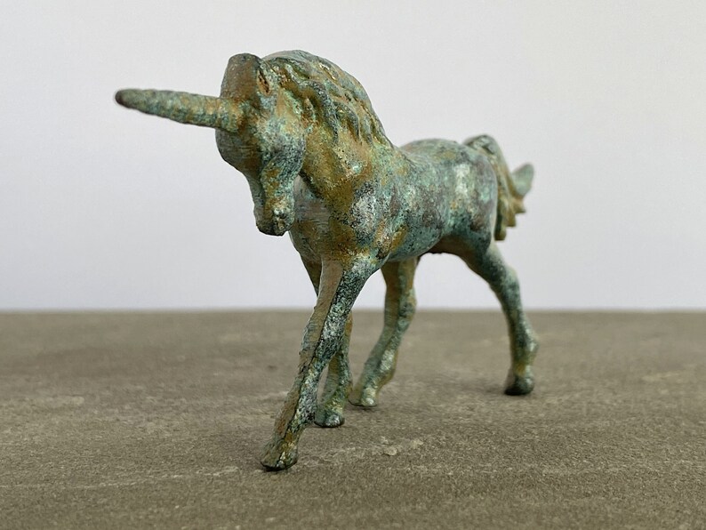 May include: A small, green and gold metal unicorn figurine. The unicorn is standing on all four legs with its head turned to the side. The unicorn has a long, curved horn.