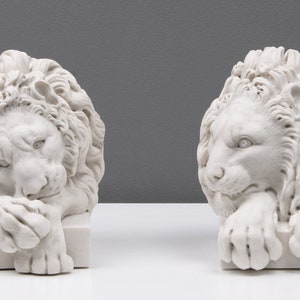 Może przedstawiać: Dwie białe marmurowe rzeźby lw&oacute;w, każda z detaliczną grzywą i spoczynkową pozą. Lwy są zwr&oacute;cone w r&oacute;żnych kierunkach, jeden patrzy w lewo, a drugi w prawo.
