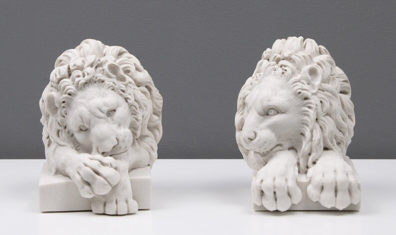 Może przedstawiać: Dwie białe marmurowe rzeźby lw&oacute;w, każda z detaliczną grzywą i spoczynkową pozą. Lwy są zwr&oacute;cone w r&oacute;żnych kierunkach, jeden patrzy w lewo, a drugi w prawo.