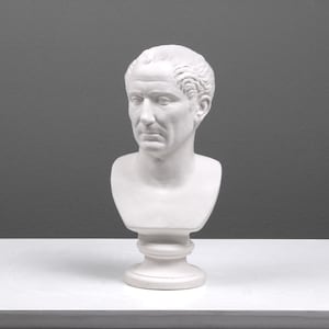 May include: White plaster bust of a man with short hair. The bust is on a white pedestal.