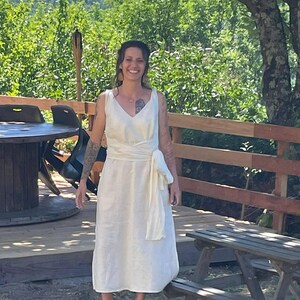 Woman wearing a sleeveless ivory wrap dress made from breathable natural linen, tied at the waist with a soft wide sash. Standing barefoot outdoors on a sunny wooden deck surrounded by lush green trees and mountain views. A relaxed, elegant look ideal for boho weddings, nature celebrations, or summer gatherings. Ships fast from the USA within 1 business day.