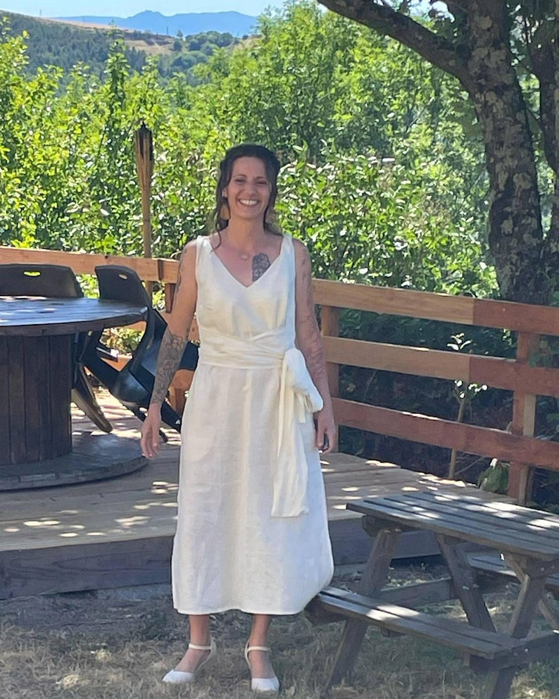 Woman wearing a sleeveless ivory wrap dress made from breathable natural linen, tied at the waist with a soft wide sash. Standing barefoot outdoors on a sunny wooden deck surrounded by lush green trees and mountain views. A relaxed, elegant look ideal for boho weddings, nature celebrations, or summer gatherings. Ships fast from the USA within 1 business day.