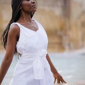 A woman in a white linen wrap dress posing by a fountain. The dress features a soft V-neckline front and back, wide twisted straps, a sewn-in waist tie for a flattering fit, and high side slits. Crafted from breathable natural linen with hidden side pockets. Ideal for romantic vacations, beach dinners, or summer events. Feminine, minimalist, and elegant style. Ships fast from the USA within 1 business day.