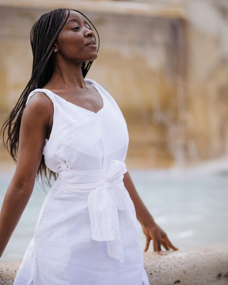 A woman in a white linen wrap dress posing by a fountain. The dress features a soft V-neckline front and back, wide twisted straps, a sewn-in waist tie for a flattering fit, and high side slits. Crafted from breathable natural linen with hidden side pockets. Ideal for romantic vacations, beach dinners, or summer events. Feminine, minimalist, and elegant style. Ships fast from the USA within 1 business day.
