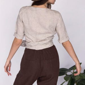 Back view of a natural beige linen wrap blouse with short cuffed sleeves, paired with high-waisted brown linen pants. The blouse ties at the waist for an adjustable fit. Made from 100% breathable linen. Ships quickly from the USA.