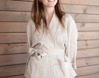 Puede incluir: Chaqueta envolvente de lino beige claro con cinturón anudado a juego y dos bolsillos delanteros. La chaqueta tiene mangas largas y cuello en V. La modelo lleva pantalones grises oscuros.