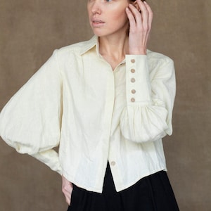 May include: A white linen blouse with puffed sleeves and a button-up front. The blouse has a relaxed fit and is cropped at the waist.