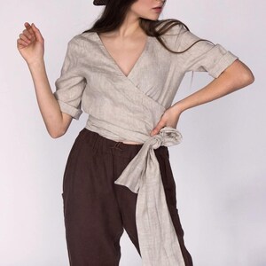 Front view of a natural beige linen wrap blouse with a deep V-neckline and hand-stitched detailing along the collar. The blouse features rolled sleeves and a long tie at the waist for an adjustable fit. Paired with relaxed brown linen trousers. Made from 100% breathable linen. Ships fast from the USA.
