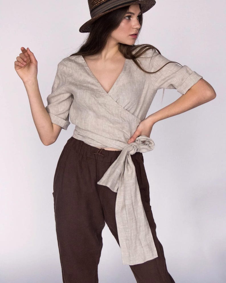 Front view of a natural beige linen wrap blouse with a deep V-neckline and hand-stitched detailing along the collar. The blouse features rolled sleeves and a long tie at the waist for an adjustable fit. Paired with relaxed brown linen trousers. Made from 100% breathable linen. Ships fast from the USA.