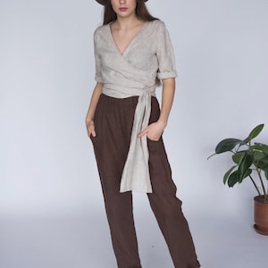 view of a woman wearing a natural linen wrap blouse with a deep V-neckline, hand-stitched collar, and long side tie for an adjustable fit. Styled with high-waisted chocolate brown linen pants and a straw hat for a relaxed summer look. Ships quickly from the USA.