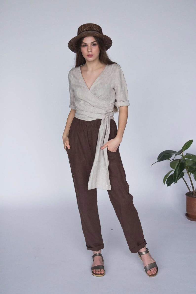 view of a woman wearing a natural linen wrap blouse with a deep V-neckline, hand-stitched collar, and long side tie for an adjustable fit. Styled with high-waisted chocolate brown linen pants and a straw hat for a relaxed summer look. Ships quickly from the USA.